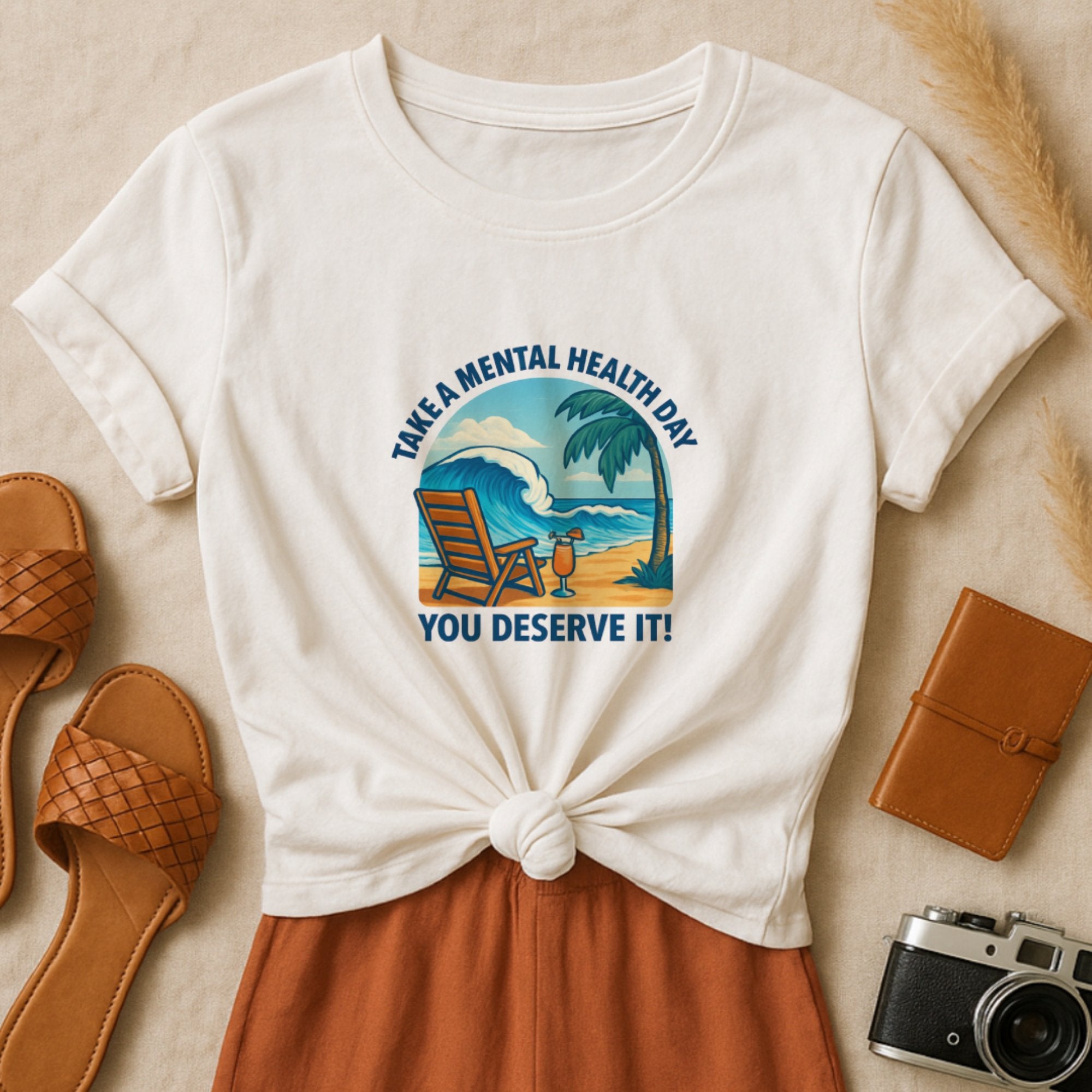 White “Take a Mental Health Day – You Deserve It” t-shirt featuring a relaxing beach scene with a lounge chair, palm tree, and ocean waves, encouraging rest, self-care, and emotional wellbeing