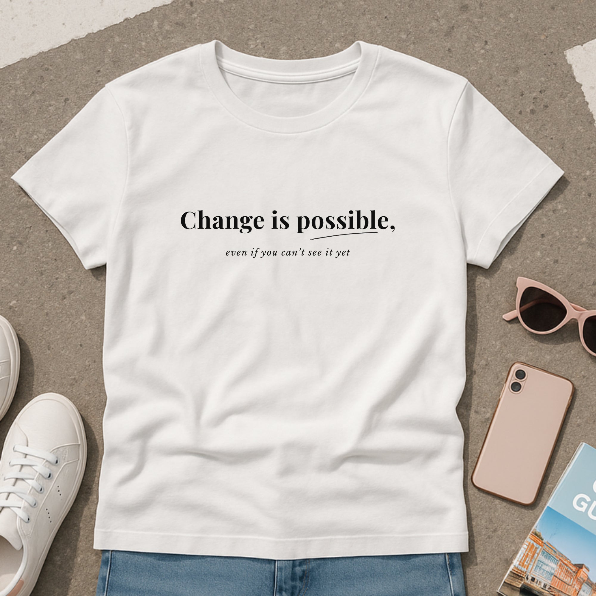 White mental health t-shirt with the message “Change is possible, even if you can’t see it yet,” styled flat lay with sneakers, sunglasses, phone, and book on a neutral concrete background