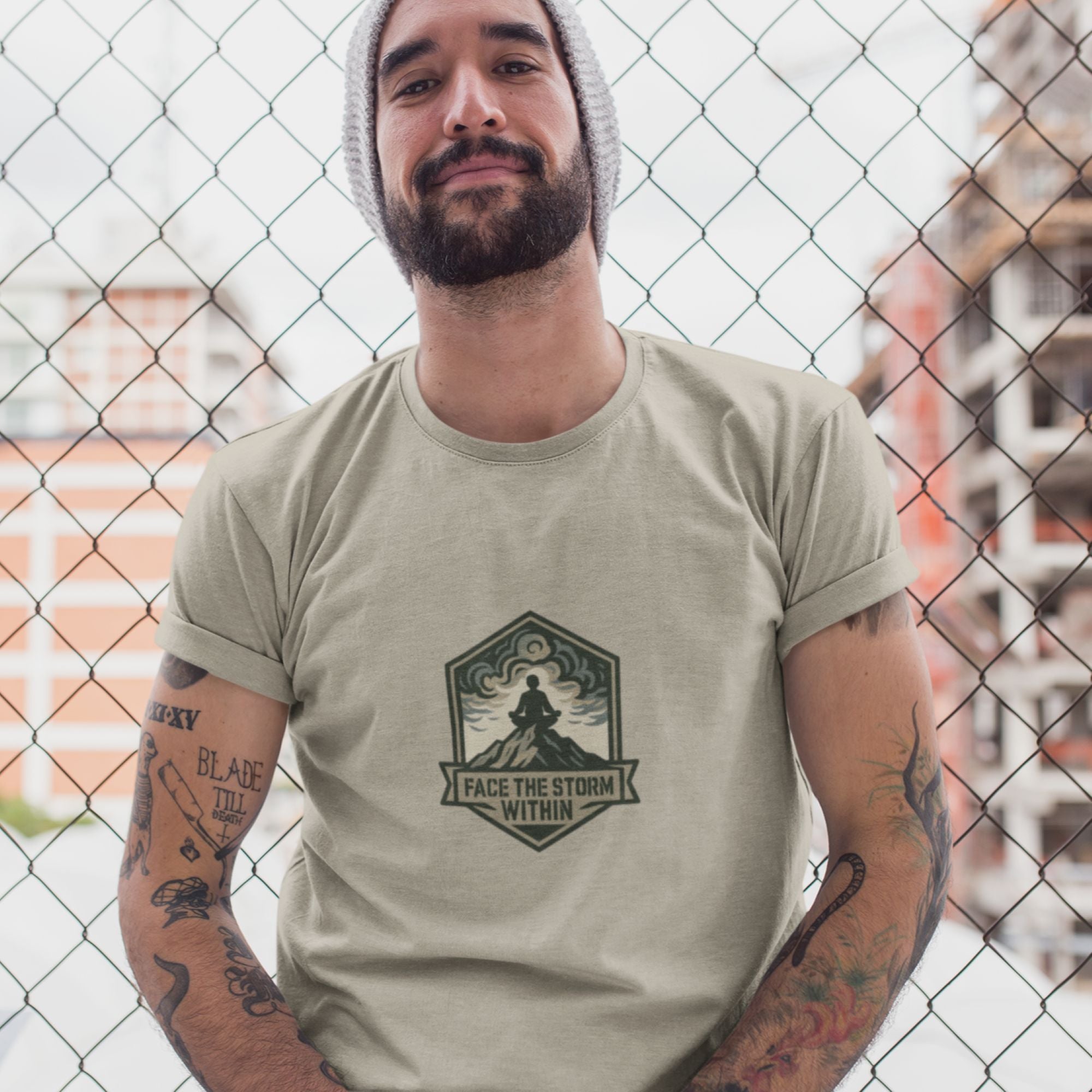 Man wearing a beige t-shirt with a graphic design and text, standing in front of a chain-link fence.