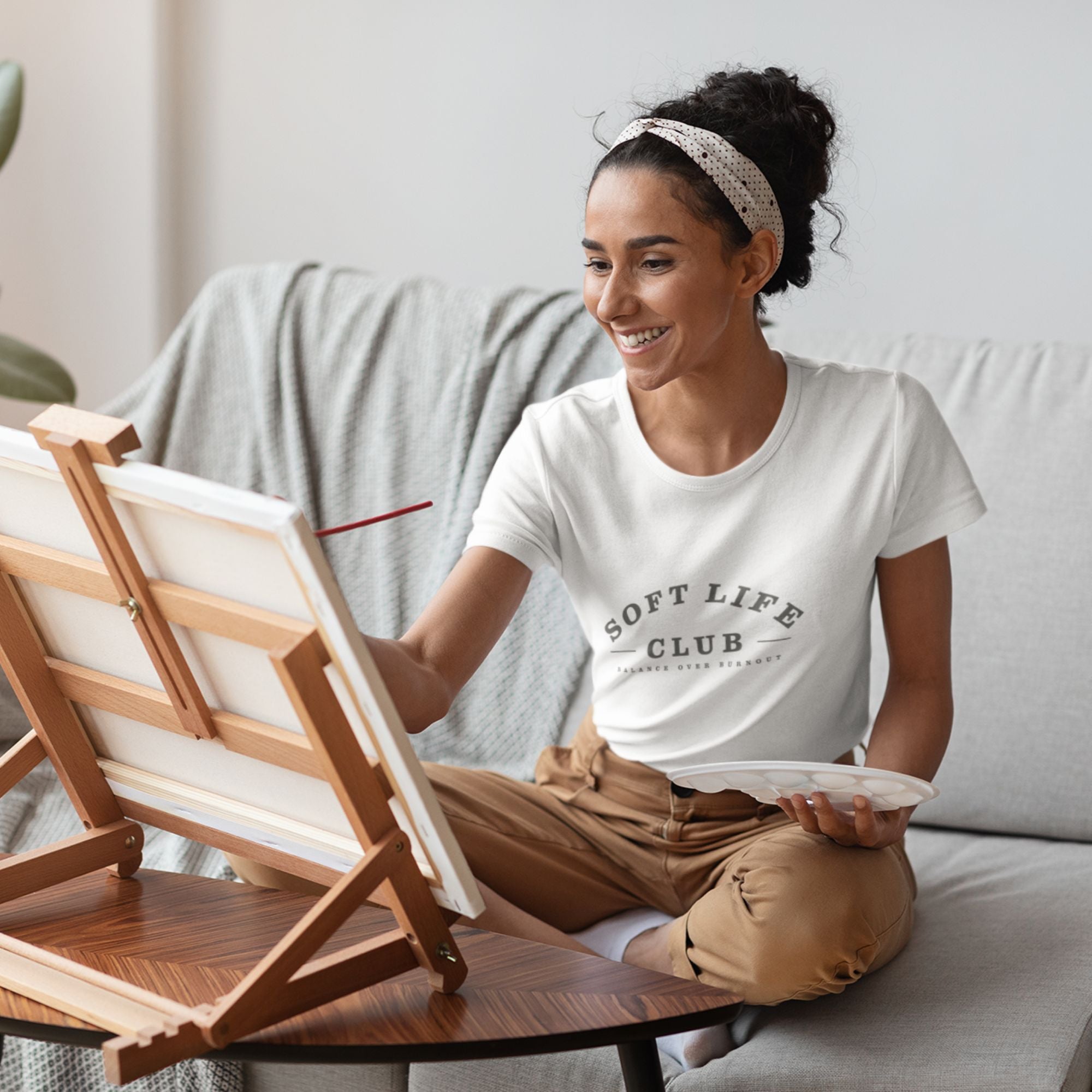 Woman sitting on a couch with a wooden easel and paintbrush, wearing a 'Soft Life Club' t-shirt.
