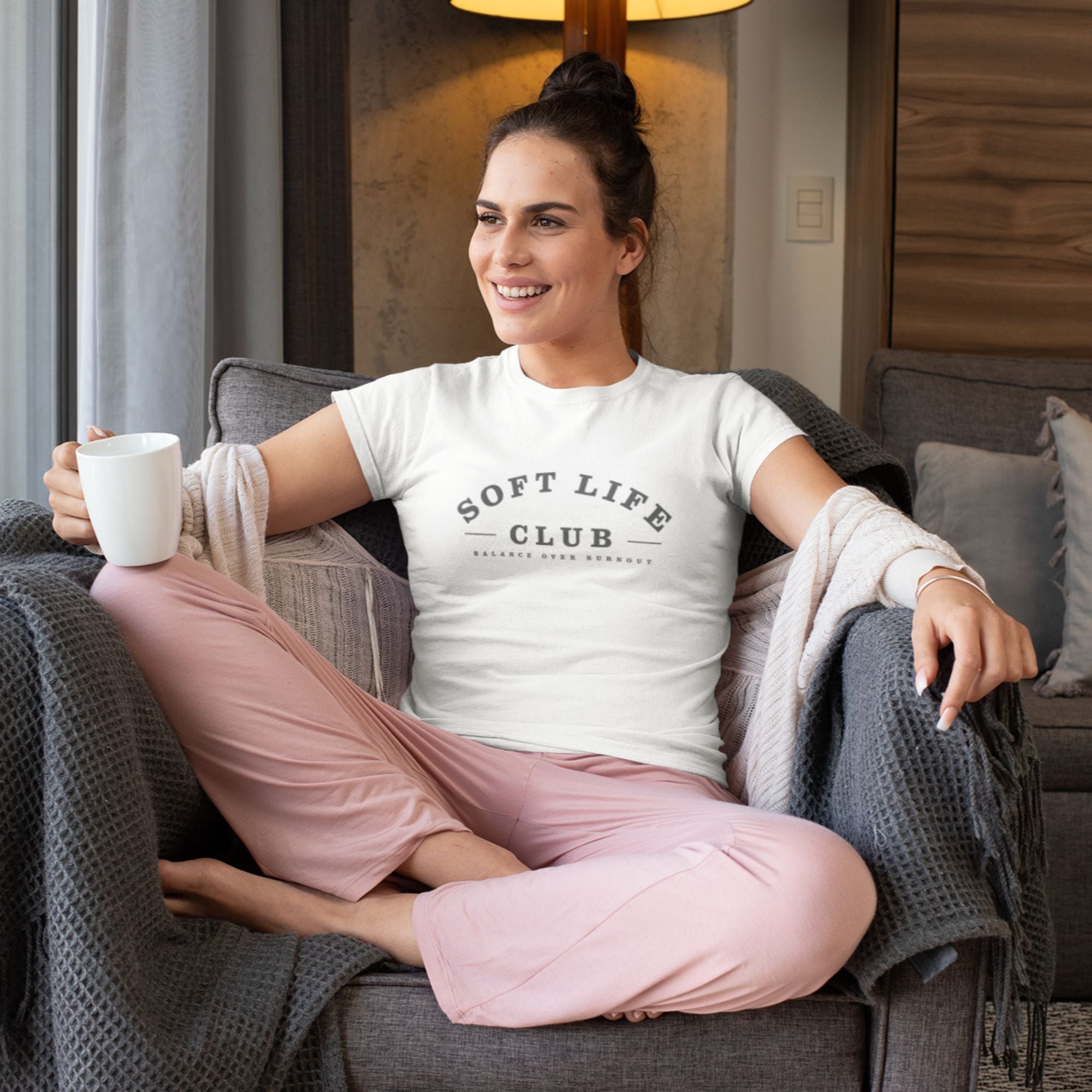 Woman sitting on a couch wearing a 'Soft Life Club' t-shirt and pink pants, holding a mug.