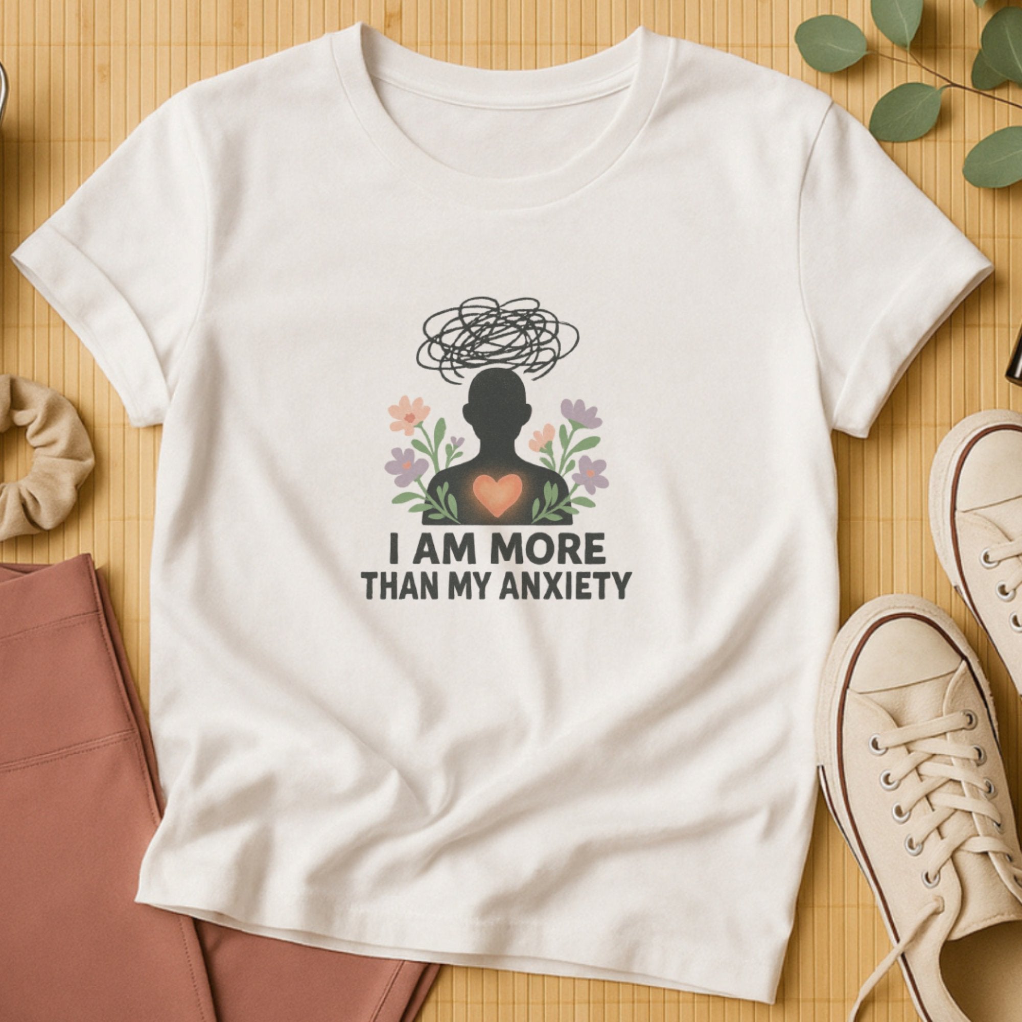White mental health affirmation t-shirt with a silhouette figure, scribble anxiety cloud, heart symbol, and flowers surrounding the phrase “I Am More Than My Anxiety,” promoting hope and self-compassion