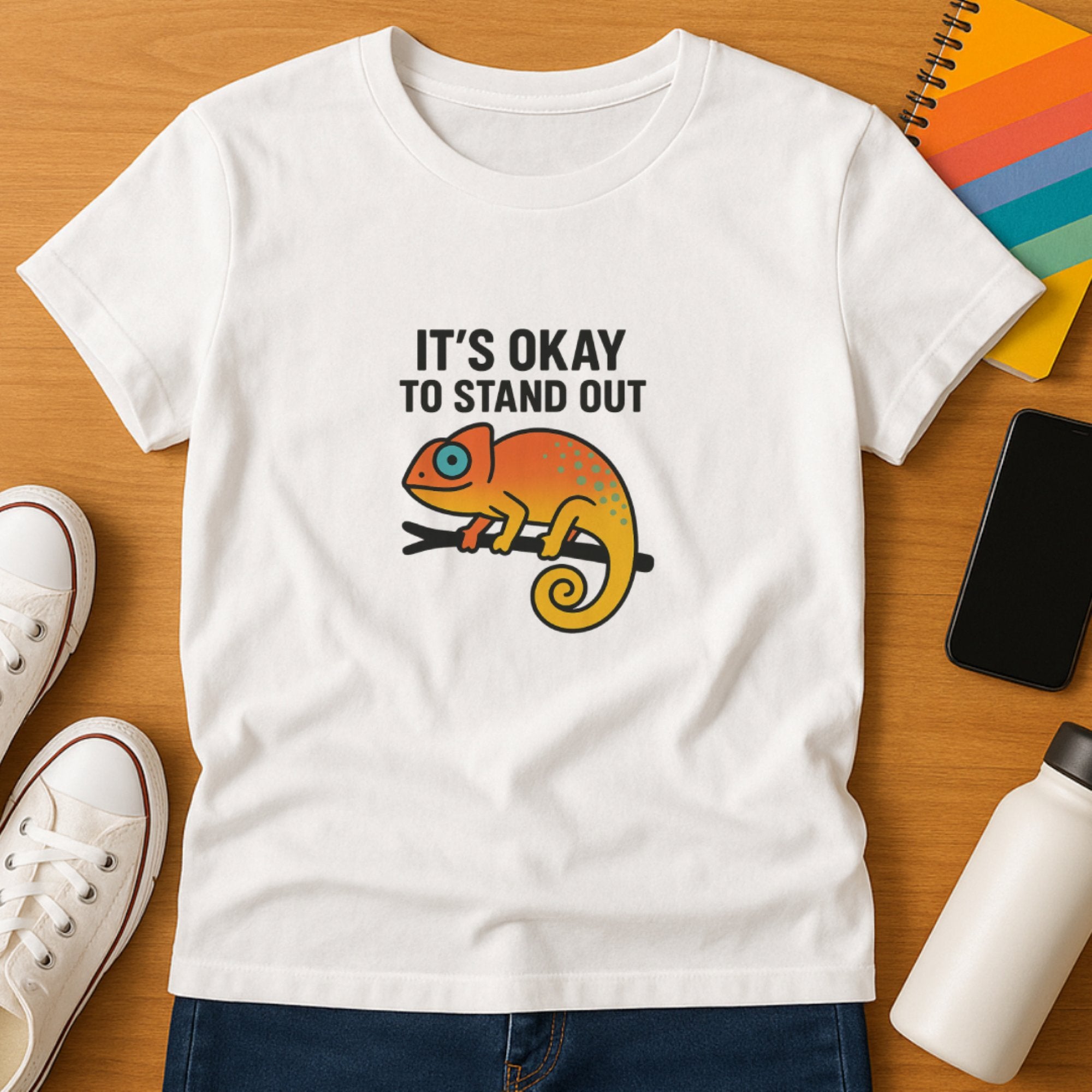 White “It’s Okay to Stand Out” t-shirt featuring a bright colorful chameleon illustration, celebrating individuality, self-acceptance, and confidence