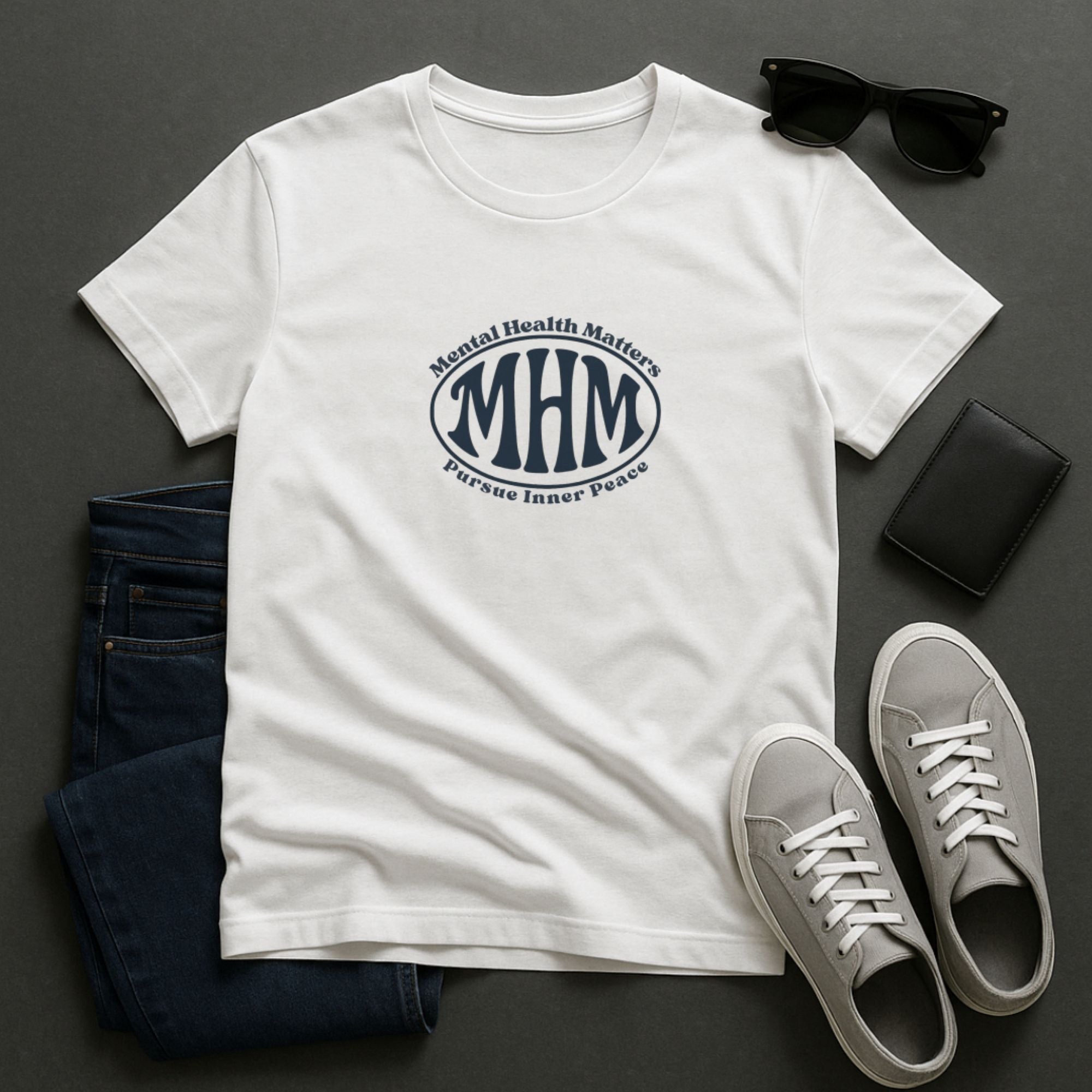 White Mental Health Matters t-shirt featuring a minimalist circular “MHM” emblem with the phrase “Pursue Inner Peace,” styled flat lay with jeans, sneakers, sunglasses, and wallet on a dark background