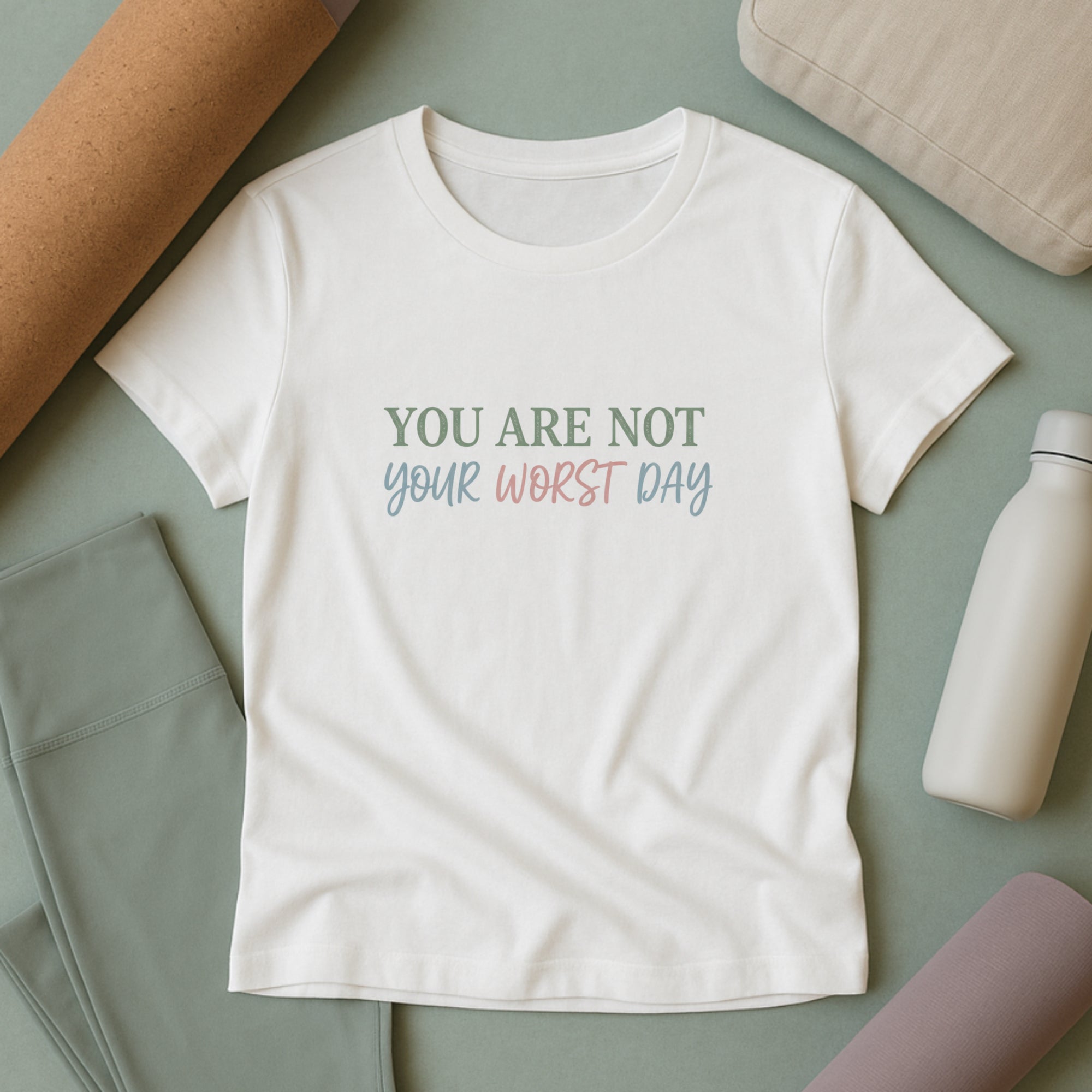 White mental health t-shirt with the phrase “You are not your worst day” in soft pastel lettering, styled flat lay with leggings, yoga mat, and water bottle on a muted green background