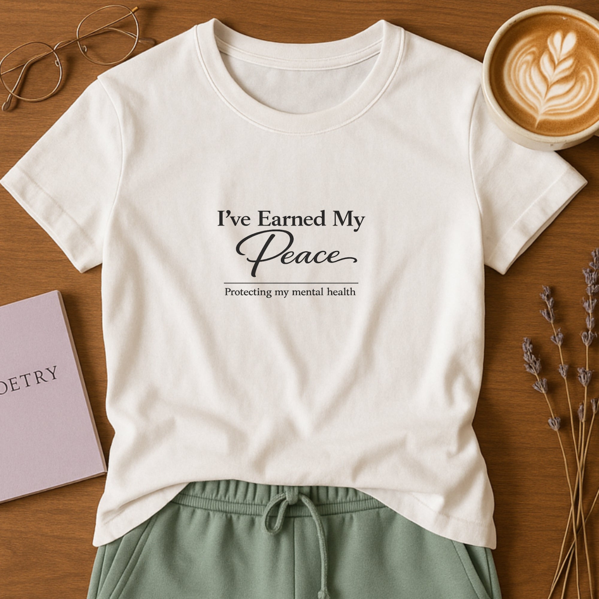 White minimalist t-shirt featuring the phrase “I’ve Earned My Peace” in elegant black typography, styled in a flat lay with coffee, book, glasses, and neutral lifestyle accents