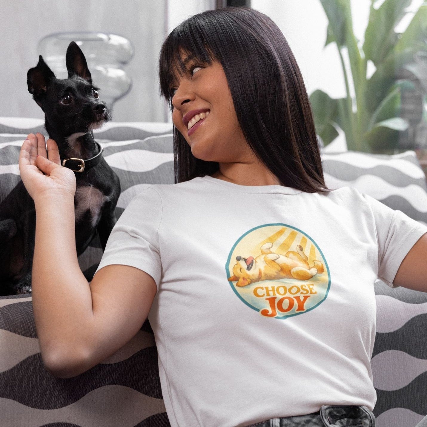 Woman wearing a white t-shirt with a “Choose Joy” dog illustration, sitting on a couch and smiling at a small black dog beside her in a cozy indoor setting