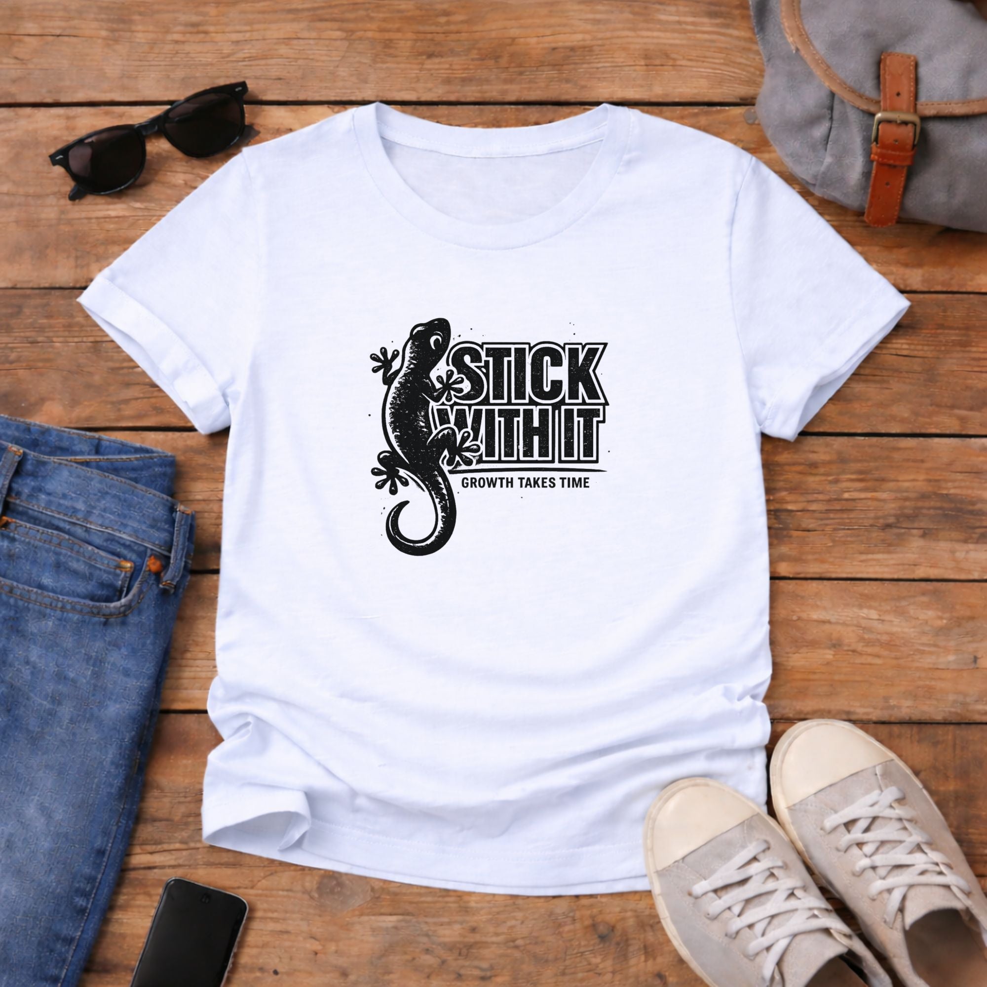 White t-shirt with black graphic and text on a wooden surface with jeans, shoes, and sunglasses.